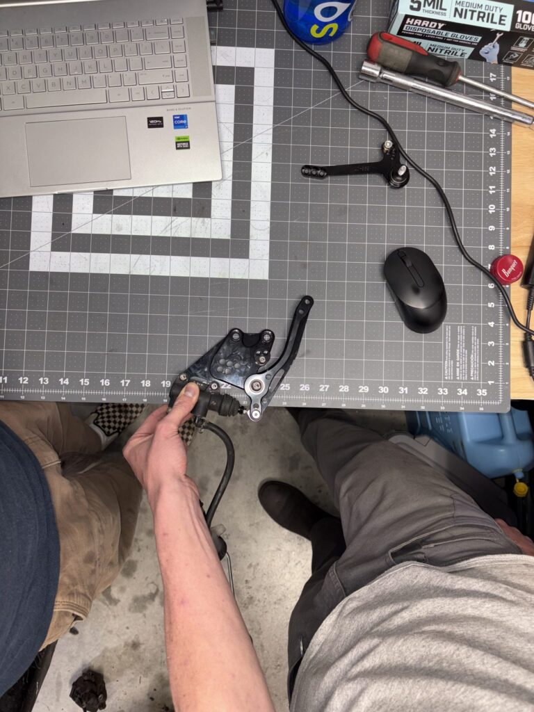 Rearset and rear brake master cylinder laid out on a cutting mat next to a laptop